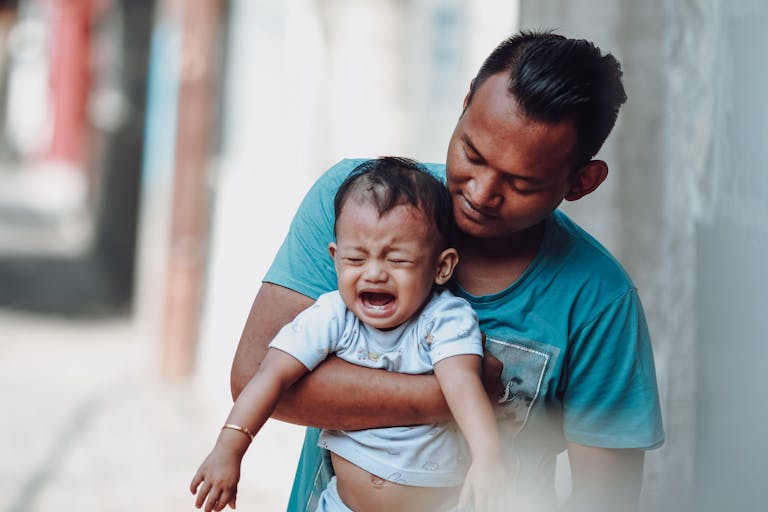 A father lovingly holds his crying baby boy outdoors, creating a tender parenting moment.